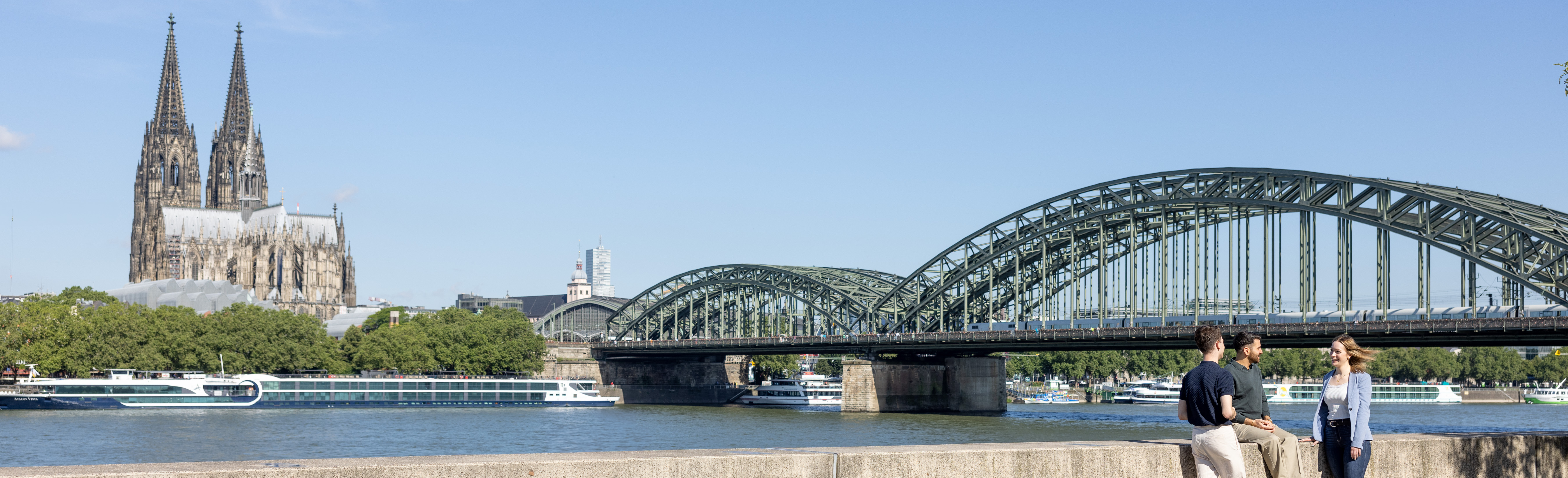 Es sind drei Personen zu sehen, die auf einer Mauer am Rheinufer sitzen. Es handelt sich um Nachwuchskräfte des LVR, die ihre Mittagspause dort verbringen.